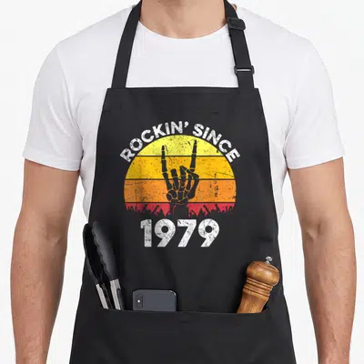 Tablier de cuisine noir avec motif vintage Rockin Since 1979 et main de rock sur fond sunset orange.