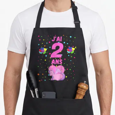 Tablier de cuisine noir avec une licorne rose, le texte J'ai 2 ans et des ballons multicolores sur fond gris uni.