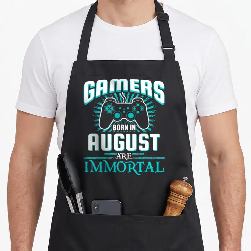 Tablier de cuisine noir avec manette de jeu vidéo et texte Gamers Born in August Are Immortal en blanc et turquoise.