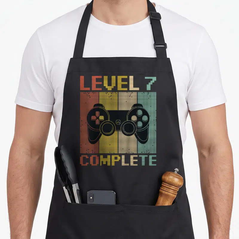 Tablier de cuisine noir avec illustration manette de jeu vidéo et texte Level 7 Complete en style rétro pixelisé multicolore.