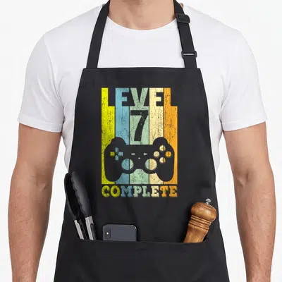 Tablier de cuisine noir avec illustration manette et texte Level 7 Complete sur fond coloré.