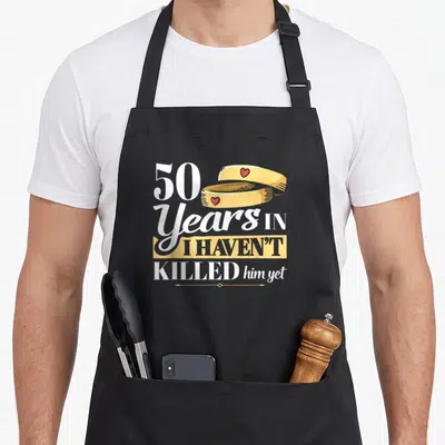 Tablier de cuisine noir avec 50 Years in I haven't killed him yet et deux anneaux dorés ornés de cœurs rouges.