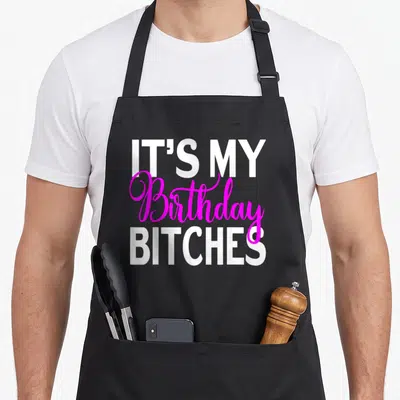 Tablier de cuisine noir avec le texte It's My anniversaire Bitches en lettres blanches et roses vives.