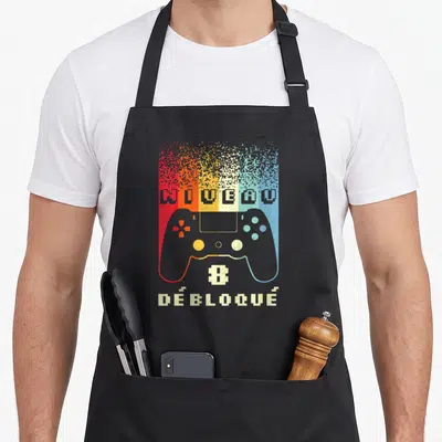 Tablier de cuisine noir avec illustration de manette de jeu vidéo et texte Niveau 8 Débloqué en style pixel art coloré.