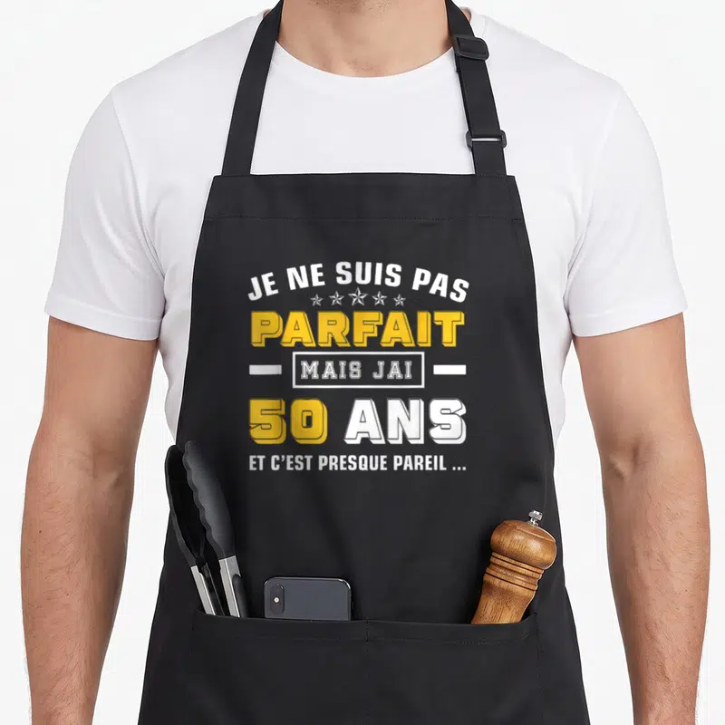 Tablier de cuisine noir avec l'inscription Je ne suis pas parfait mais j'ai 50 ans en lettres jaunes et blanches.