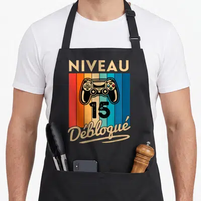 Tablier de cuisine noir avec manette de jeu, le chiffre 15 et le mot Débloqué sur fond rayé multicolore.