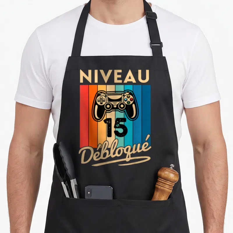 Tablier de cuisine noir avec manette de jeu, le chiffre 15 et le mot Débloqué sur fond rayé multicolore.