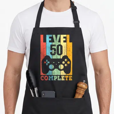 Tablier de cuisine noir avec inscription Level 50 Complete et silhouette de manette sur fond de bandes verticales colorées délavées.