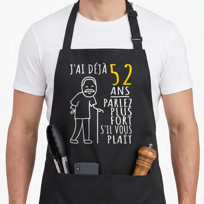 Tablier de cuisine noir avec une illustration d'homme barbu au bâton et le texte J'ai déjà 52 ans parlez plus fort s'il vous plaît en blanc et jaune.