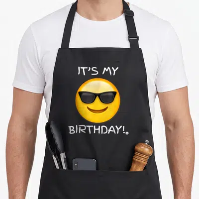 Tablier de cuisine noir avec un emoji jaune à lunettes noires et texte blanc celebrating l'anniversaire