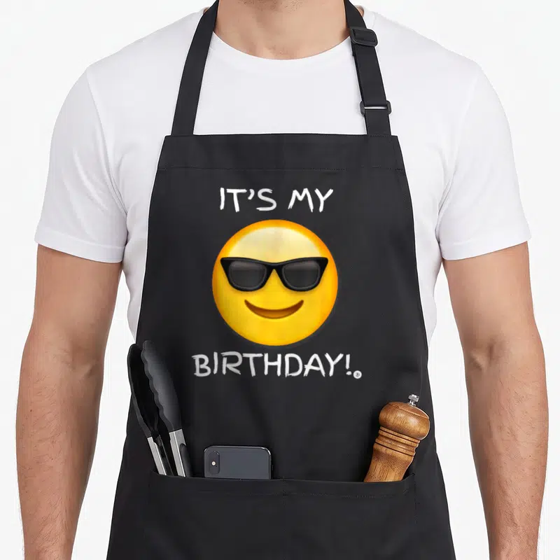 Tablier de cuisine noir avec un emoji jaune à lunettes noires et texte blanc celebrating l'anniversaire