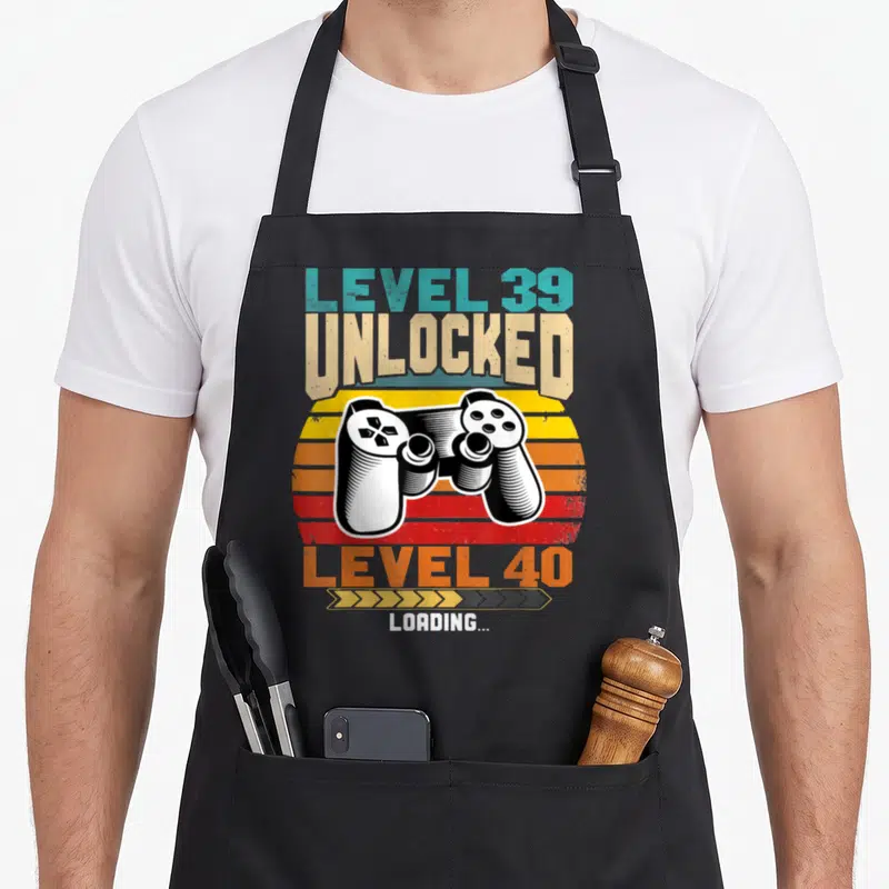 Tablier de cuisine noir avec manette de jeu vidéo et texte Niveau 39 Unlocked Level 40 Loading sur fond de soleil rétro coloré.
