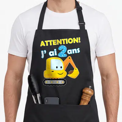 Tablier de cuisine noir avec un excavateur jaune souriant et le texte Attention j'ai 2 ans en lettres colorées.