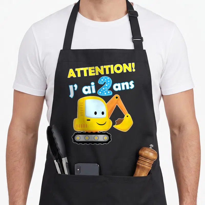 Tablier de cuisine noir avec un excavateur jaune souriant et le texte Attention j'ai 2 ans en lettres colorées.