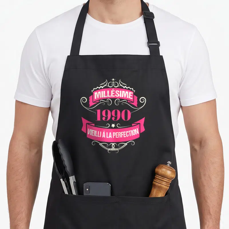 Tablier de cuisine noir avec une étiquette rose et blanche Millésime 1990 Vieilli à la perfection.