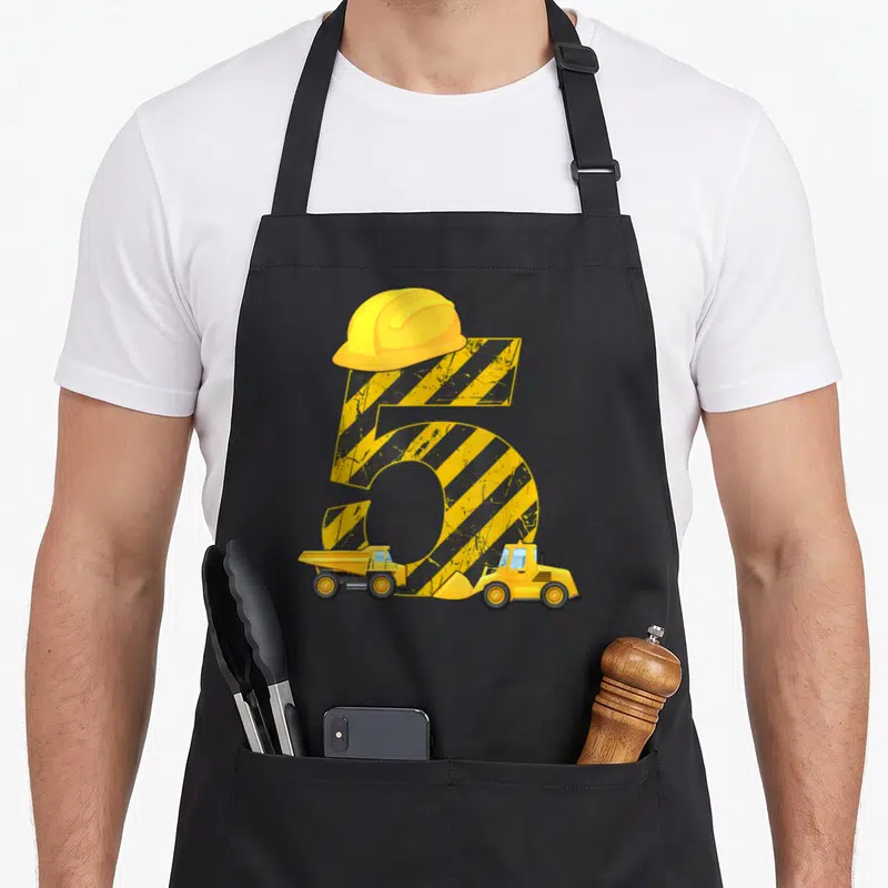 Tablier de cuisine noir avec un chiffre 5 jaune et noir surmonté d'un casque, accompagné d'un excavateur et d'un camion benne.
