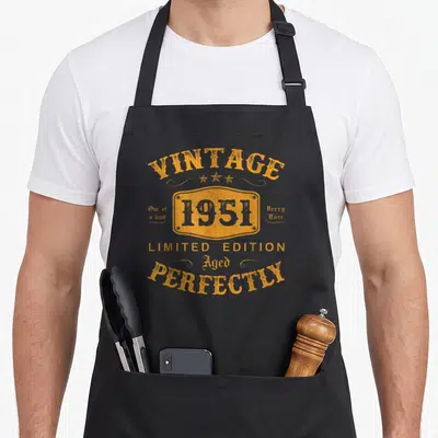 Tablier de cuisine noir avec inscription vintage orange 1951 Aged Perfectly Limited Edition et trois étoiles décoratives au centre.