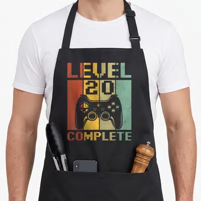 Tablier de cuisine noir avec illustration manette de jeu vidéo et texte Level 20 Complete en style rétro.