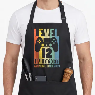 Tablier de cuisine noir avec illustration manette de jeu et texte Niveau 12 débloqué couleur vintage.