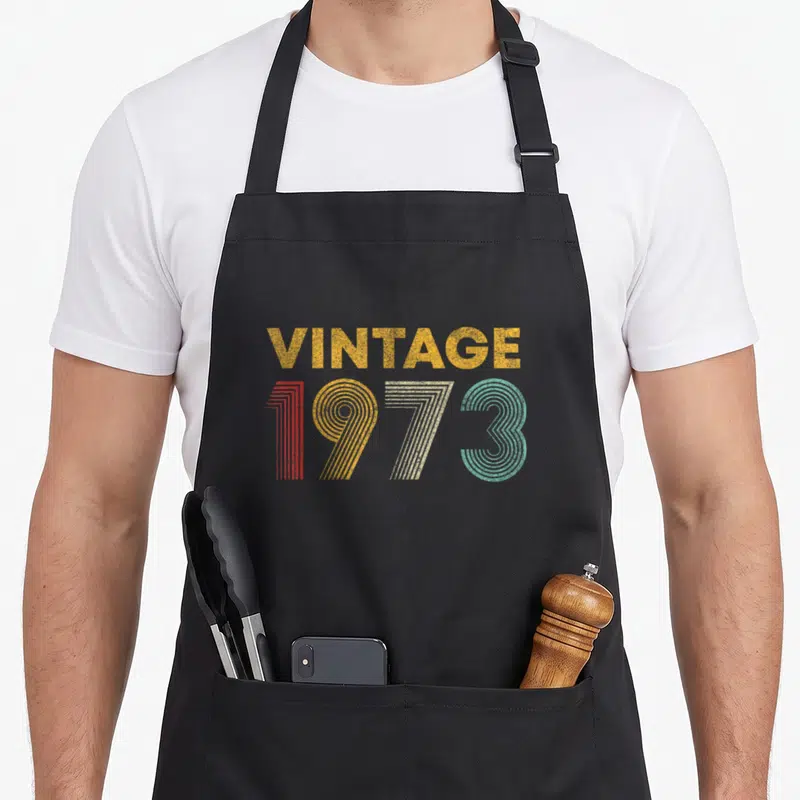 Tablier de cuisine noir avec motif vintage 1973 aux couleurs rétro et texte doré texturé.