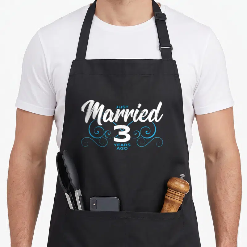 Tablier de cuisine noir avec l'inscription Just Married 3 Years Ago en blanc et des volutes décoratives bleues.
