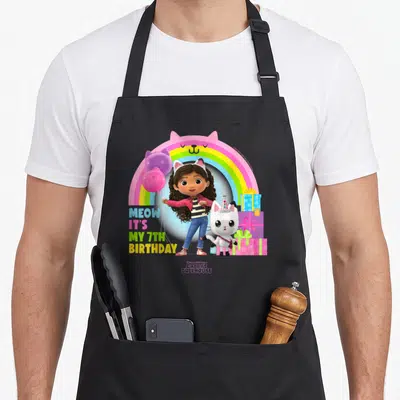 Tablier de cuisine noir avec Gabby, son chat blanc et l'inscription Meow It's My 7th anniversaire sous un arc-en-ciel rose.
