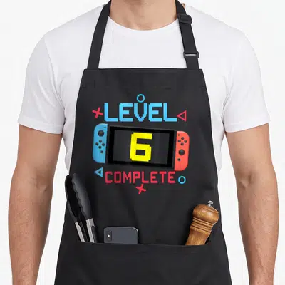 Tablier de cuisine noir avec une illustration de console de jeu et le texte Level 6 Complete en lettres pixelisées colorées.