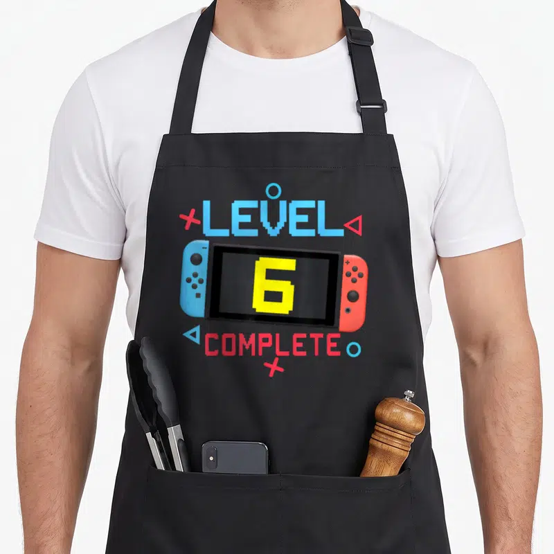 Tablier de cuisine noir avec une illustration de console de jeu et le texte Level 6 Complete en lettres pixelisées colorées.