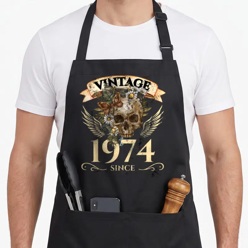 Tablier de cuisine noir avec une tête de mort vintage 1974 ornée de fleurs blanches et de papillon.