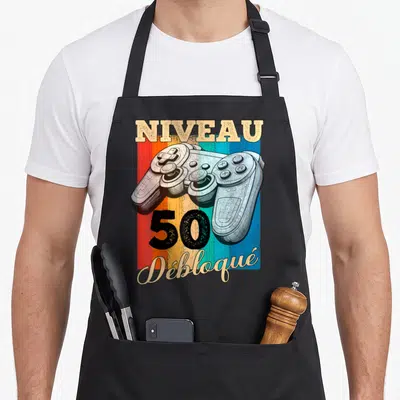 Tablier de cuisine noir avec illustration vintage manette de jeu et texte Niveau 50 Débloqué sur fond coloré.
