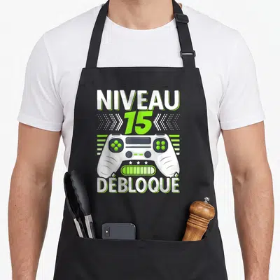 Tablier de cuisine noir avec illustration manette de jeu vidéo et texte Niveau 15 Débloqué en blanc et vert lime.