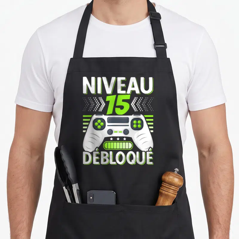 Tablier de cuisine noir avec illustration manette de jeu vidéo et texte Niveau 15 Débloqué en blanc et vert lime.