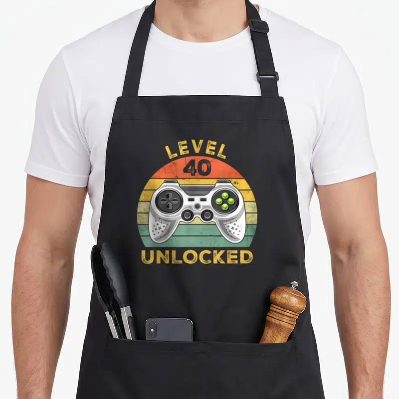 Tablier de cuisine noir avec illustration manette de jeu vidéo et texte Level 40 Unlocked sur fond arc-en-ciel vintage.