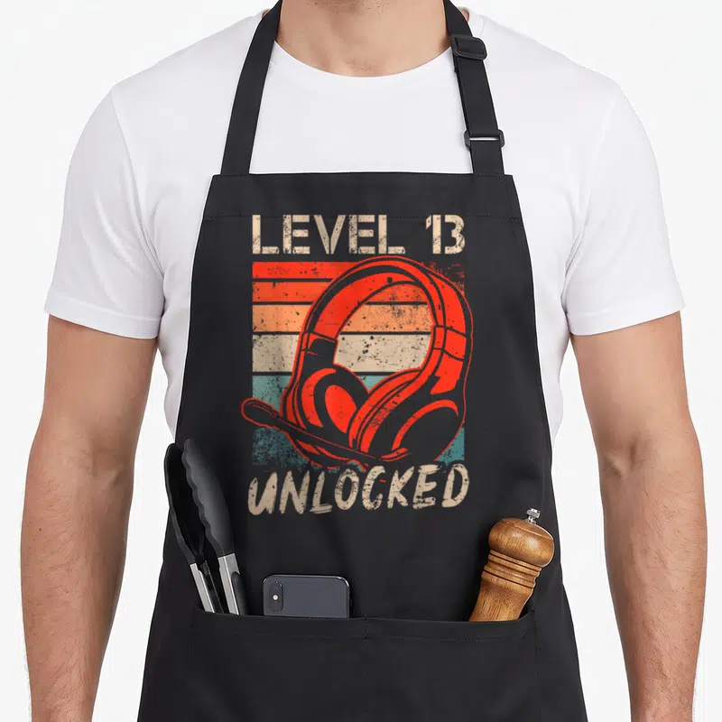 Tablier de cuisine noir avec illustration casque rouge et texte Level 13 Unlocked style vintage.