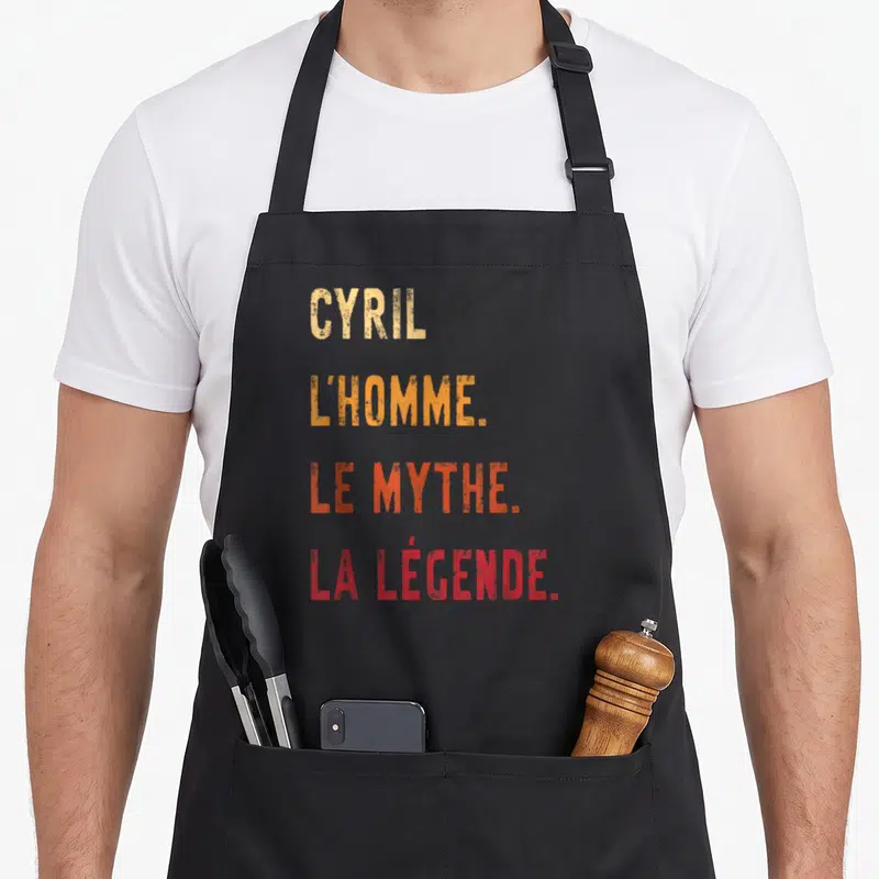 Tablier de cuisine noir avec inscription Cyril l'homme le mythe la légende en lettres dégradées orange et rouge.
