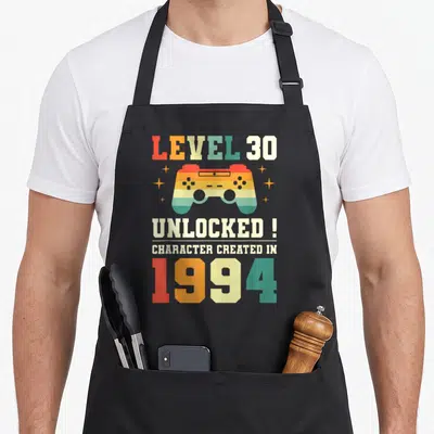Tablier de cuisine noir avec une manette de jeu colorée et le texte Level 30 UnlockedCharacter Created in 1994 imprimé au centre.