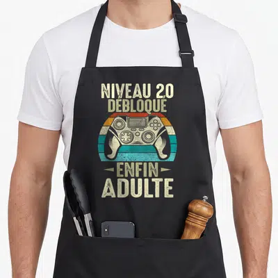 Tablier de cuisine noir avec illustration vintage manette de jeu vidéo et texte Niveau 20 Débloque Enfin Adulte imprimé en lettres capitales.