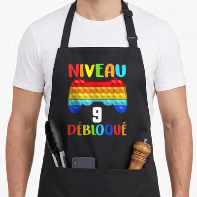 Tablier de cuisine noir avec un pop it gamepad multicolore et le texte Niveau Débloqué 9 ans imprimé au centre.