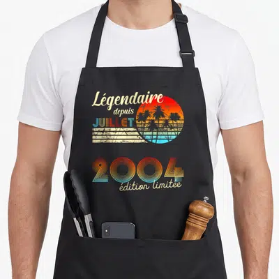 Tablier de cuisine noir avec design rétro soleil couchant palmiers et texte Légendaire depuis Juillet 2004