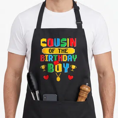 Tablier de cuisine noir avec inscription pixelisée multicolore et trophées dorés sur fond uni.