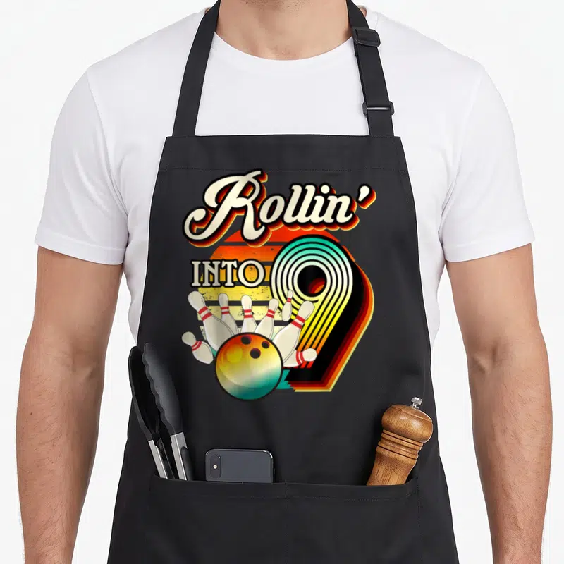 Tablier de cuisine noir avec illustration bowling rétro et texte Rollin' Into 9 coloré.