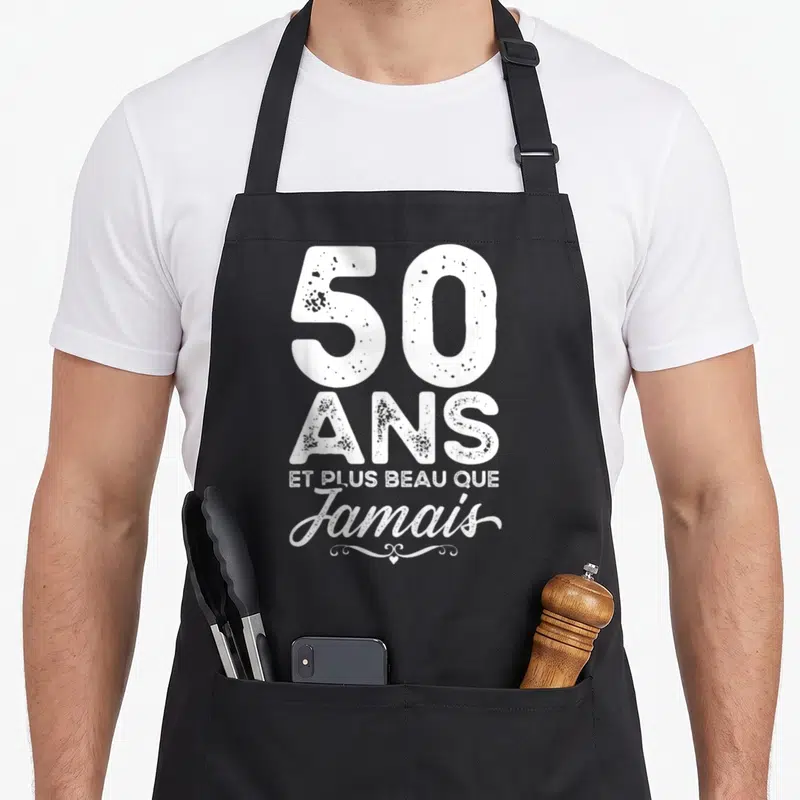 Tablier de cuisine noir avec inscription 50 ans et plus beau que jamais en typographie blanche vintage.