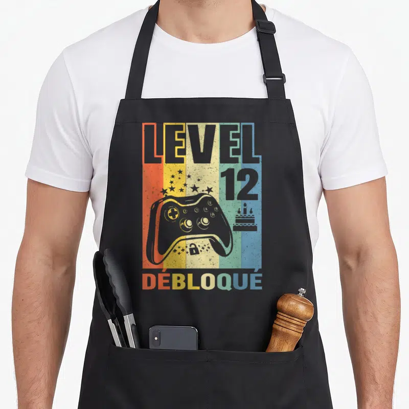 Tablier de cuisine noir avec illustration manette de jeu vidéo Level 12 Débloqué sur fond rayé coloré.