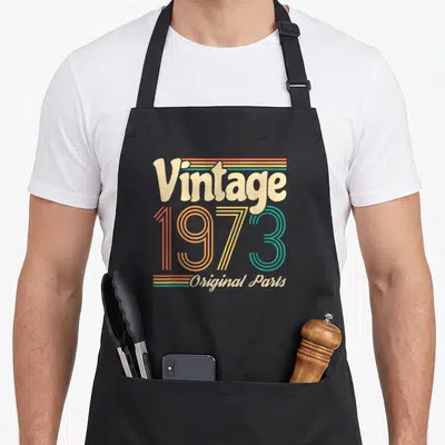 Tablier de cuisine noir avec le motif vintage 1973 Original Parts en couleurs pastel et rayures horizontales.
