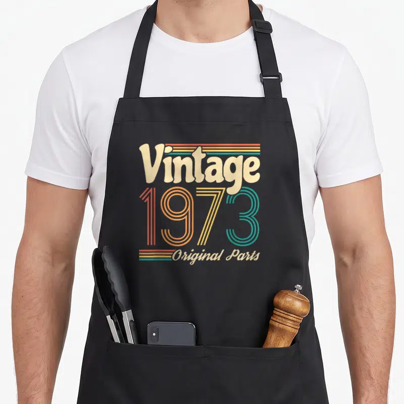 Tablier de cuisine noir avec le motif vintage 1973 Original Parts en couleurs pastel et rayures horizontales.