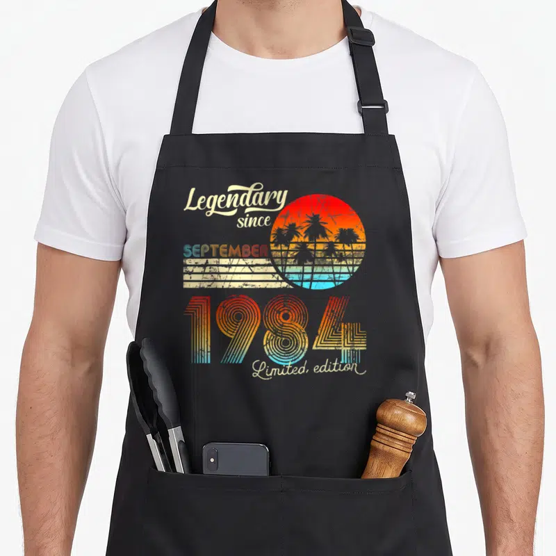 Tablier de cuisine noir avec design vintage coloré affichant 1984 et un soleil couchant aux palmiers.