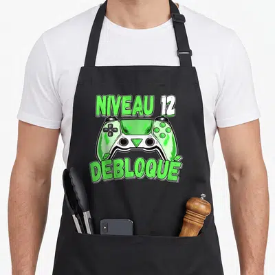 Tablier de cuisine noir avec illustration manette verte et texte Niveau 12 Débloqué en typographie stylisée.