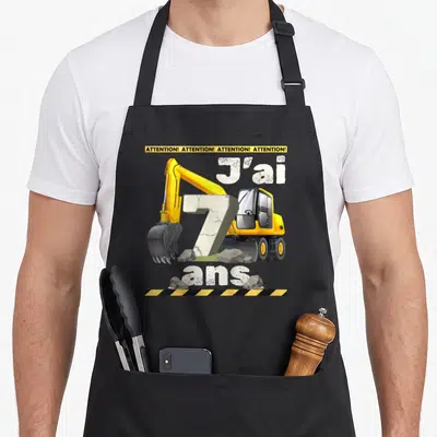 Tablier de cuisine noir avec illustration de pelleteuse jaune et texte J'ai 7 ans sur fond de gravats.