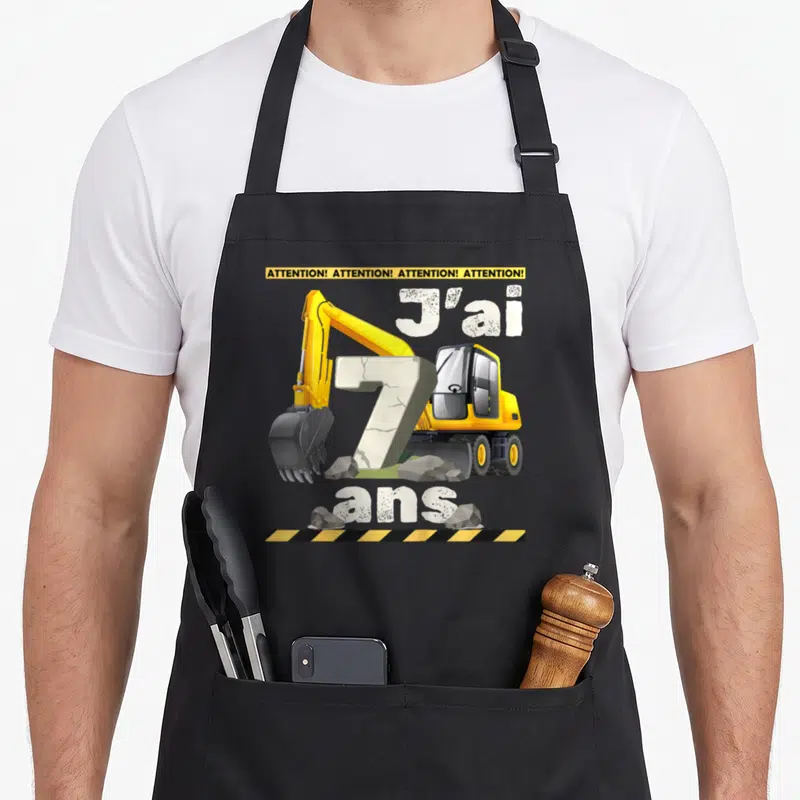 Tablier de cuisine noir avec illustration de pelleteuse jaune et texte J'ai 7 ans sur fond de gravats.