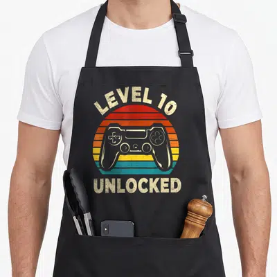 Tablier de cuisine noir avec illustration vintage manette de jeu vidéo et texte Level 10 Unlocked en lettres blanches usées.